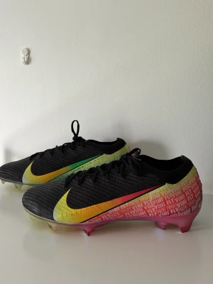 Nike Mercurial, VINI VOA VINI FLY (limited edition) - Snygga Nike Mercurial fotbollsskor med svart ovandel, stora färgskiftande swoosh-loggor i gult, grönt och rött samt textdetaljer på sidorna. Sulan har en regnbågsskimrande finish och dobbar för bra grepp på planen. Lätta och smidiga för snabba rörelser.