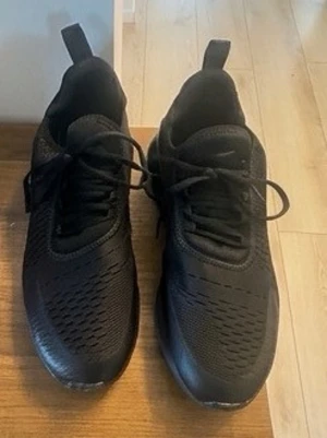 Svarta Nike Air Max sneakers - Snygga svarta Nike Air Max sneakers med mesh-ovandel och kraftig synlig Air Max-sula. Skorna har lågt skaft, klassisk snörning och diskret logga på plösen. Perfekta för en sportig och stilren look.