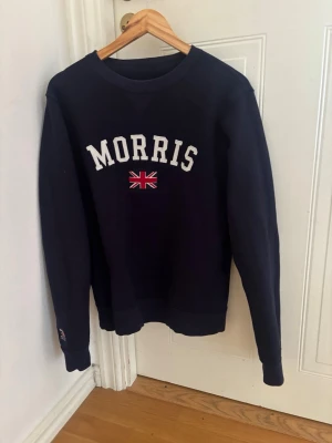 Mörkblå sweatshirt från Morris - Mörkblå sweatshirt från Morris med vit broderad logga och brittisk flagga på bröstet. Klassisk rund halsringning och ribbade muddar vid ärmslut och nederkant. Perfekt för en stilren och avslappnad look.