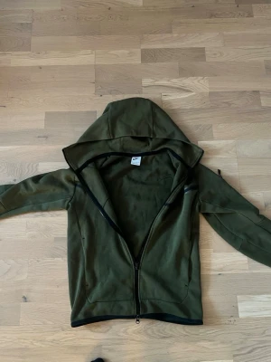 Nike tech - Snygg olivgrön hoodie från Nike med hel dragkedja framtill och svart detalj över bröstet. Tröjan har huva, två sidofickor och svarta kantband vid ärmslut och nederkant. Perfekt för en avslappnad och sportig stil.