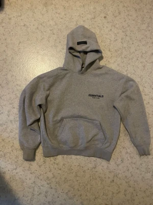Grå Essentials Fear of God hoodie - Snygg grå hoodie från Essentials Fear of God med svart logga på bröstet och stor text på ryggen. Klassisk känguruficka framtill och ribbade muddar. Perfekt oversized passform och mjukt material som känns riktigt skönt mot huden.