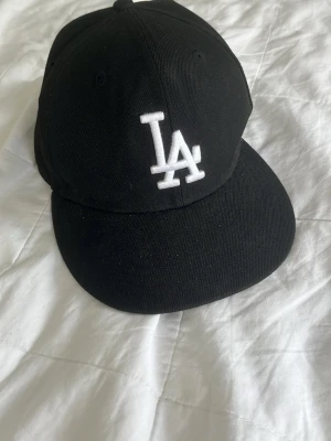 Svart LA-keps från New Era - Snygg svart keps från New Era med LA-logga broderad i vitt framtill. Klassisk modell med böjd skärm och ventilerande hål upptill. Perfekt accessoar för en sportig och urban stil.