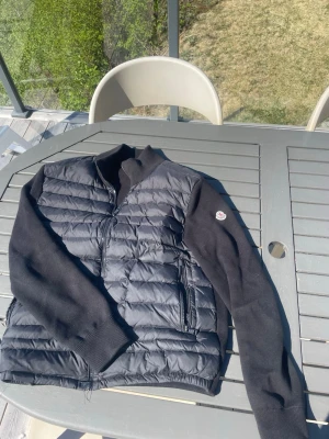 Svart cardigan från Moncler - Snygg svart Cardigan från Moncler med quiltad front och rygg i glansigt material samt stickade ärmar. Jackan har två dragkedjefickor och en hög krage. Moncler-logga på vänster ärm. Perfekt för kalla dagar.