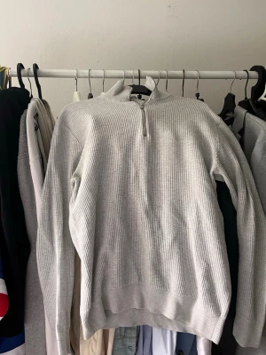 Ljusgrå halvzip tröja - Ljusgrå ribbstickad tröja från brothers med halv zip i halsen. Tröjan har lång ärm och en avslappnad passform, perfekt för lager-på-lager. Materialet är mjukt och ger en clean look.