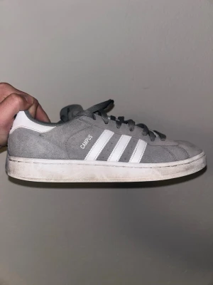 Grå Adidas Campus sneakers - Säljer ett par grå Adidas Campus sneakers i mocka med klassiska vita ränder på sidan och vit sula. Skorna har lågt skaft och snörning framtill. Perfekta för dig som gillar stilrena och tidlösa sneakers.