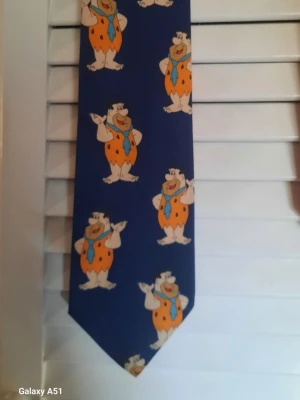 Blå slips med Fred Flinta-motiv - Rolig blå slips med motiv av Fred Flinta från Familjen Flinta. Perfekt för dig som gillar tecknade serier och vill sticka ut med en färgglad accessoar. Slipsen har en klassisk form och är tillverkad i ett slätt tyg.  Original från Hanna & Barbara