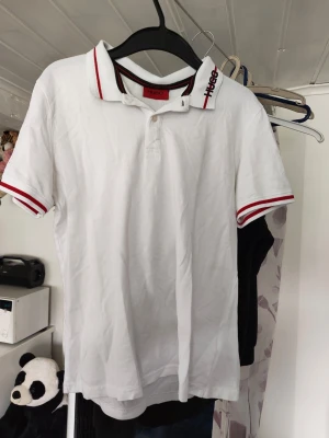 Vit pikétröja från Hugo Boss - Vit pikétröja från Hugo Boss med röda detaljer på krage och ärmslut. Klassisk polokrage med knappar och logga broderad på kragen. Perfekt för en stilren och sportig look. Finns någon fläck vet ej om de går bort Kan inte garantera äkthet 