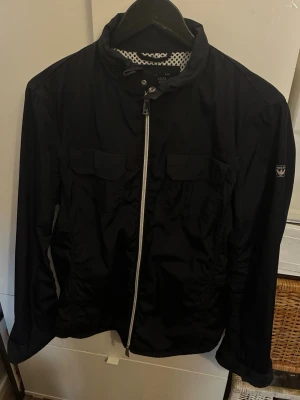 Svart vindjacka från Armani Jeans - Svart vindjacka från Armani Jeans med vit dragkedja, två bröstfickor med lock och meshfoder. Jackan har hög krage med tryckknapp och en sportig, stilren look. Perfekt för blåsiga dagar och enkel att matcha med olika outfits.