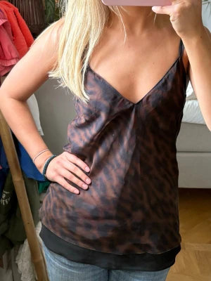 Leopardmönstrad linnetopp med v-ringning - Snygg leopardmönstrad linnetopp i bruna och svarta toner med v-ringning och tunna axelband. Toppen har en lös passform och är perfekt för en trendig look. Materialet känns lätt och svalt mot huden.