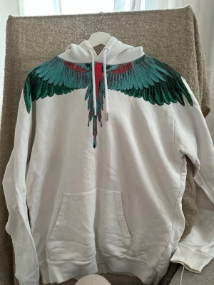 Marcel Burlon hoodie - Säljer en vit hoodie från Marcelo Burlon med ikoniskt vingtryck i grönt, blått och rött över axlar och bröst. Hoodien har lite tecken på missfärgning runt kragen. Nypris 4500kr. 