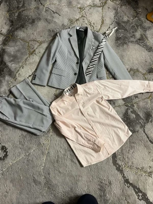 Randig grå kostym med skjorta - Snygg grå kostym med diskreta ränder, kavaj och matchande byxor. Ingår gör en ljusrosa skjorta och en randig slips. Perfekt för dig som vill ha en stilren och modern look. Kavajen har klassisk krage och knappar framtill.