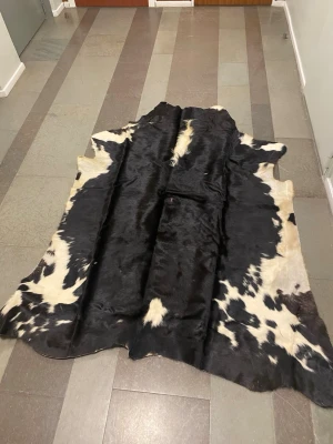 Svartvit kohudsmatta använd i nyskick, ca 190x215cm - Säljer en stor kohudsmatta i svart och vitt med naturligt mönster. Mattan har en mjuk och glansig yta och passar perfekt som inredningsdetalj i hemmet. Materialet är äkta kohud och formen är oregelbunden, vilket ger en unik känsla. Längre hår på vissa platser. Nypris 7500kr. Bäst hämta i Stor Stockholm.