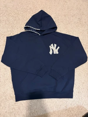 Mörkblå New York Yankees Custom hoodie - Snygg mörkblå hoodie med stor NY Yankees-logga i beige brodyr på bröstet. Huva med vit dekorationssöm och klassisk känguruficka framtill. Perfekt för dig som gillar sportig streetstyle och vill sticka ut med en ikonisk look.