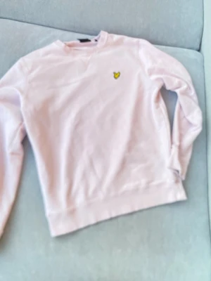 Ljusrosa sweatshirt från Lyle & Scott - Säljer en ljusrosa sweatshirt från Lyle & Scott med deras klassiska gula fågel-logga broderad på bröstet. Tröjan har rund halsringning, långa ärmar och är gjord i mjukt bomullsmaterial. Perfekt för en avslappnad och stilren look.