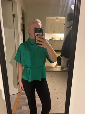 Grön topp från H&M - Snygg grön blus med hög krage och volang nedtill. Blusen har korta, vida ärmar och ett skärp i midjan som ger en markerad siluett. Storlek M!