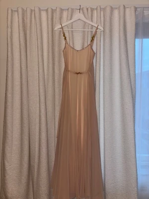 Ljus puderrosa långklänning Ida Sjöstedt - Elegant långklänning i puderrosa från Ida Sjöstedt. Klänningen har tunna axelband med guldfärgade Ida Sjöstedt-detaljer och ett matchande bälte i midjan. Tillverkad i ett lätt, flytande material som ger en lyxig känsla.