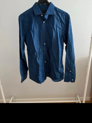 Blå mönstrad skjorta med knappar - Säljer en blå skjorta med diskret prickigt mönster och klassisk krage. Skjortan har långa ärmar och vita knappar framtill. Perfekt för dig som gillar stilrena detaljer och en modern look.