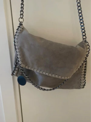 Grå stella mcartney liknande axelväska med kedjedetaljer - Grå axelväska i mocka med silverfärgade kedjor längs kanterna och som axelrem. Väskan har ett lock och dekorativ söm runt hela väskan. Perfekt för dig som gillar en stilren men edgy look.