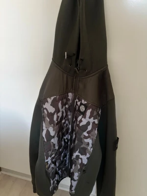 Stone Island hoodie med camomönster - Säljer en grön hoodie från Stone Island i storlek M. Framsidan har ett grått och vitt camomönster och dragkedja hela vägen. Klassisk Stone Island-patch på bröstet och justerbar huva med snörning. Materialet är en mix av bomull och syntet för extra komfort.