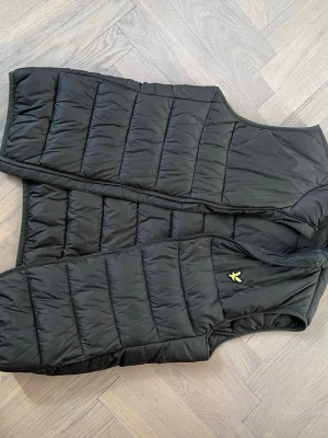 Mörkgrön  dunväst från Lyle & Scott - Snygg svart dunväst från Lyle & Scott med det klassiska gula logomärket på bröstet. Västen har quiltad design, hög krage och dragkedja framtill. Perfekt att slänga över en hoodie eller tröja för extra värme.