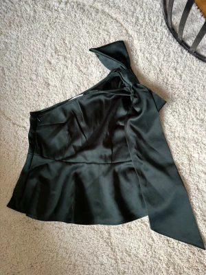 Mörkgrön oneshoulder topp med rosett - !!Prislappen är kvar!!Stilren mörkgrön oneshoulder topp i satinliknande material med stor rosett på axeln. Aldrig använd då den var för liten