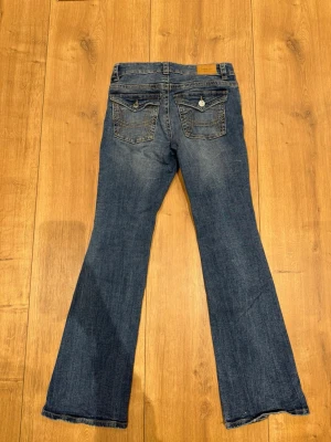 Blå bootcut jeans från Gina Tricot - Säljer ett par blå bootcut jeans från Gina Tricot med slitningar på framsidan och klassiska bakfickor med knappdetaljer. Jeansen har normal passform och är tillverkade i bomull med en snygg tvättad look.