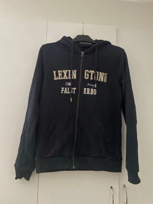 Marinblå zip-hoodie från Lexington - Snygg marinblå hoodie från Lexington med dragkedja framtill och broderad text på bröstet. Den är i väldigt bra skick inga defekter o så. 
