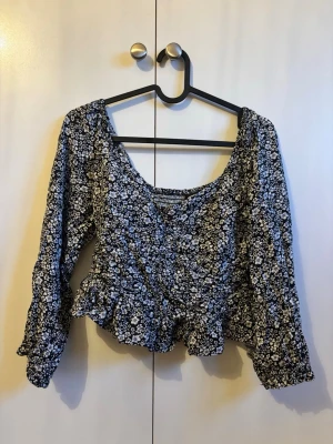 Blommig croppad topp med volang - Söt svart croppad topp med vitt blommönster och volangkant. Toppen har långärmade puffärmar och en djupare ringning framtill. 