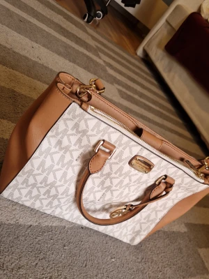  Michael Kors Kellan Satchel - Snygg handväska från Michael Kors i beige och ljusbrunt konstläder med MK-logga över hela väskan. Väskan har guldfärgade metalldetaljer, dubbla handtag och dragkedja upptill. Perfekt för dig som gillar klassisk och trendig design.  Pris kan diskuteras vid snabb affär.