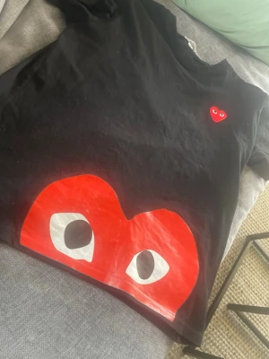 Svart Comme des Garçons Play t-shirt - Svart t-shirt från Comme des Garçons Play med ikoniskt rött hjärta med ögon på bröstet och ett stort hjärta med ögon tryckt nedtill. Klassisk passform och rund hals. Perfekt för dig som gillar streetwear och vill sticka ut. Storlek M men passar bäst som S