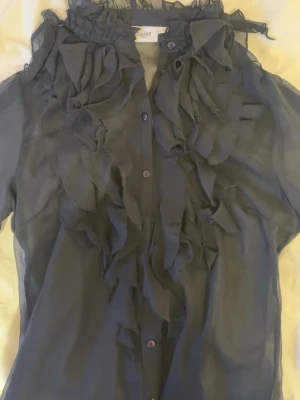 Svart volangblus från Saint Tropez - Snygg svart blus från Saint Tropez med transparent tyg och volangdetaljer framtill. Blusen har knäppning hela vägen och en hög krage med volanger. Perfekt för dig som vill ha en statement piece i garderoben.