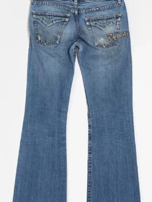 Blå bootcut jeans från Replay - Säljer ett par blå bootcut jeans från Replay med slitningar och broderad logga på bakfickan. Jeansen har låg midja, utsvängda ben och är tillverkade i denim. Perfekta för dig som gillar retrovibbar.
