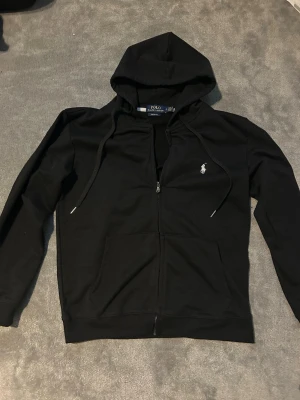 Svart hoodie från Polo Ralph Lauren - Snygg svart hoodie från Polo Ralph Lauren med dragkedja framtill och klassisk vit logga broderad på bröstet. Hoodien har huva med dragsko och två fickor framtill. Tillverkad i mjukt bomullsmaterial som är skönt att bära.