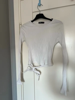Vit ribbad croppad topp - Säljer en vit ribbad croppad topp med långa ärmar och knytband i midjan. Toppen har en rund halsringning och är tillverkad i ett mjukt, stretchigt material som sitter snyggt på kroppen. Perfekt för att styla med höga jeans eller kjol.