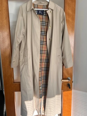 Beige trenchcoat från Burberry - Burberry trenchcoat med klassiskt nova check-foder. Lång modell med knappar framtill, två fickor och detaljerade manschetter med knapp. Tillverkad i bomullsmix. Öppning i bak som går att stänga igen med knappar om man vill det💗