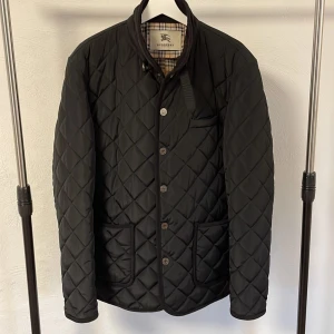 quiltad jacka från Burberry - Säljer en stilren svart vintage quiltad jacka från Burberry. Riktigt snygg och perfekt att köra lagar på lager med! Jackan har en klassisk design med knappar framtill och en diskret krage. Insidan har det ikoniska Burberry-mönstret. Perfekt för en elegant look. Pris kan såklart diskuteras!Perfekt jacka för vintern om man vill va lite finare!