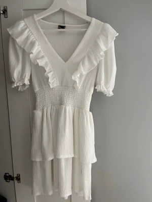 Vit volangklänning Gina Tricot - Supersöt vit kortklänning från Gina Tricot med v-ringning, volangdetaljer och puffärmar. Klänningen har smock i midjan och är tillverkad i ett lätt, strukturerat tyg som ger en luftig känsla. Perfekt för sommarens alla tillfällen! Säljer den pågrund av att jag bara fått användning av den en gång på Konformation.