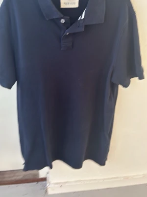 Mörkblå pikétröja från Peak Performance - Klassisk mörkblå pikétröja från Peak Performance med krage och knappar framtill. Tillverkad i mjuk bomull och har korta ärmar. Perfekt för en avslappnad och stilren look. Storlek S men passar även M