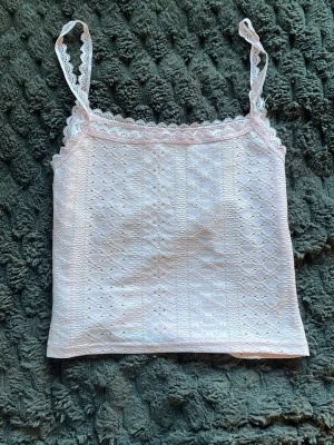 Ljusrosa spetslinnetopp - Söt ljusrosa linnetopp med spetsdetaljer längs axelband och urringning. Toppen har ett broderat hålmönster över hela tyget och smala axelband. Perfekt för en trendig och feminin look.