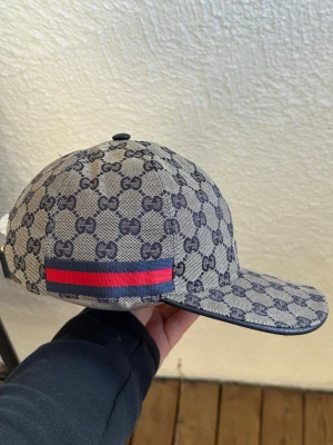 Gucci keps med GG-mönster - Säljer en snygg keps från Gucci med klassiskt GG-monogram i mörkblått på beige botten. Kepsen har en böjd skärm och en ikonisk röd-blå rand på sidan. Tillverkad i canvasmaterial som ger en exklusiv känsla och streetwear-vibe.