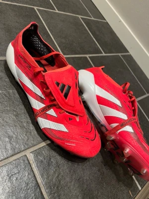 Adidas Predator röda fotbollsskor - Snygga röda Adidas Predator fotbollsskor med vita och svarta detaljer. Skorna har klassisk snörning med täckande plös och mönstrad ovandel för bättre bollkontroll. Yttersulan har fasta dobbar för spel på gräs. Perfekta för dig som vill sticka ut på planen.
