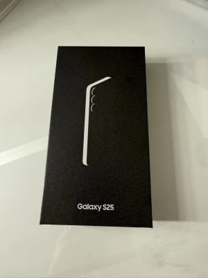 Samsung Galaxy S25 mobiltelefon - Säljer en Samsung Galaxy S25 i originalförpackning. Lådan är svart med stilren design och tydlig Galaxy S25-logga. Perfekt för dig som vill ha en modern smartphone med snygg look och avancerade funktioner. Materialet på förpackningen är kartong.