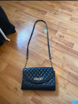 Svart quiltad axelväska med kedja - Snygg svart axelväska i quiltat mönster med guldfärgad kedjedetalj på locket och axelrem med kedja. Väskan har en klassisk rektangulär form och stängs med lock. Perfekt för dig som gillar trendiga accessoarer med lite lyxig känsla.