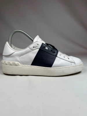 Valentino Garavani vita sneakers - Snygga vita sneakers från Valentino Garavani med en bred mörkblå remsa över mitten och coola nitar på hälen. Tillverkade i läder med klassisk låg profil och vita snören. Perfekta för dig som vill ha en stilren men ändå unik look. Storlek 37 men passar 38