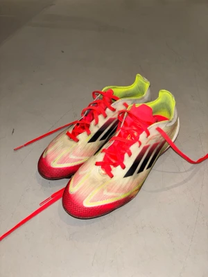 Adidas F50 Fibertouch Röd Vit - Snygga fotbollsskor från Adidas med neonröda snören och detaljer, svart logga på sidan och insida i gul neon. Ovandelen är i syntetmaterial med en strömlinjeformad design för snabbhet på planen. Perfekta för dig som vill sticka ut på fotbollsplanen.