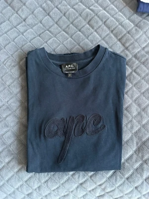 Mörkblå t-shirt från A.P.C. - Snygg mörkblå t-shirt från A.P.C. med ett stort broderat 'apc'-motiv på bröstet. Klassisk rund halsringning och mjukt bomullsmaterial som känns skönt mot huden. Perfekt för en clean och stilren look. Perfekt inför sommaren och väldigt bra skick. Skirv vid mer info eller mer bilder. Kan inte vissa hur den sitter på!!
