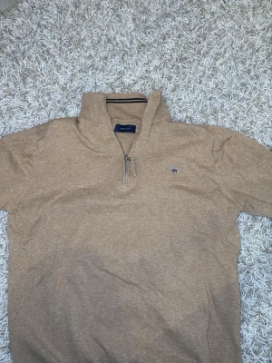 Beige stickad halvzip från Gant - Snygg beige stickad tröja från Gant med halvzip och hög krage. Tröjan har broderad Gant-logga på bröstet och ribbade detaljer vid krage och ärmslut. Perfekt för lager på lager och en clean look.