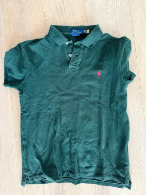 Grön piké från Polo Ralph Lauren - Säljer en klassisk mörkgrön polotopp från Polo Ralph Lauren med korta ärmar och krage. Toppens ikoniska rosa logga är broderad på bröstet och den har knappar framtill. Perfekt för dig som gillar stilrena och tidlösa plagg.