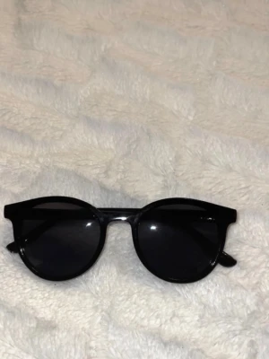 Svarta runda solglasögon - Snygga svarta solglasögon med rundad form och bred båge. Klassisk och stilren design som passar till de flesta outfits. Glasen är mörka och ramen är i plast.