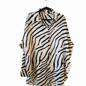 Oversized zebramönstrad skjorta - Snygg oversized skjorta med svartvitt zebramönster. Skjortan har klassisk krage, knäppning framtill och långa ärmar. Perfekt för dig som vill sticka ut med ett djurinspirerat plagg. Luftigt material och avslappnad passform.